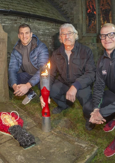 Night photo of three men crouched at grave with wreath and flaming torch.