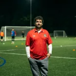 Le fondateur du club, Delwyn Derrick, debout sur un terrain de football, tandis qu'un match se déroule en arrière-plan.
