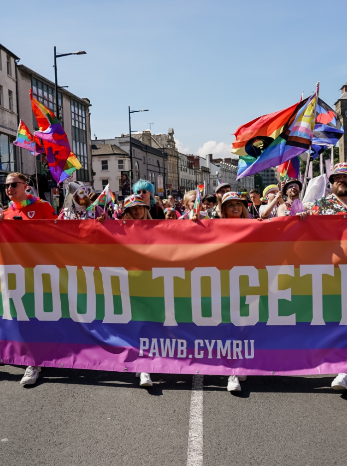 A group of LGBTQIA+ supporters carrying a rainbow banner that says Proud Together