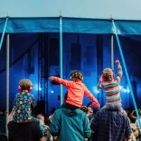 Three children sat on the shoulders of their parents listening to a band playing at a festival.