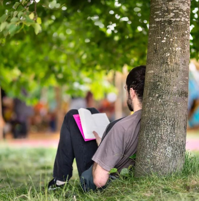 A person leaning against a tree in a park reading a book.