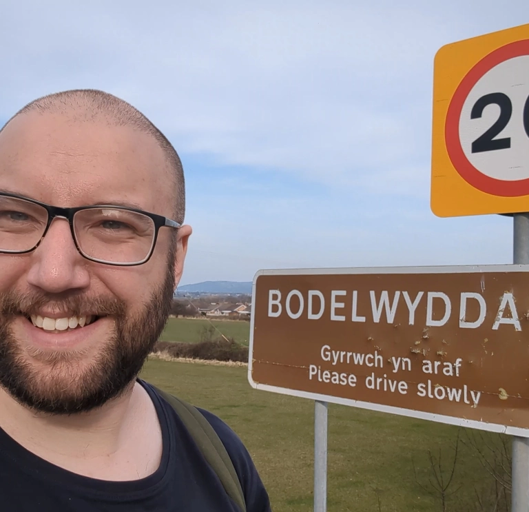 Joe taking a selfie beside a “Bodelwyddan” village sign in a grassy landscape.