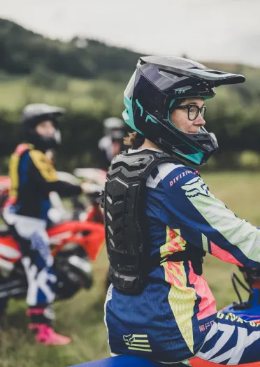 A person wearing a helmet and motorbiking safety gear, holding a motorbike and smiling
