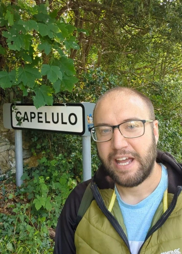 Joe standing in front of a “Capelulo” village sign, surrounded by dense greenery.