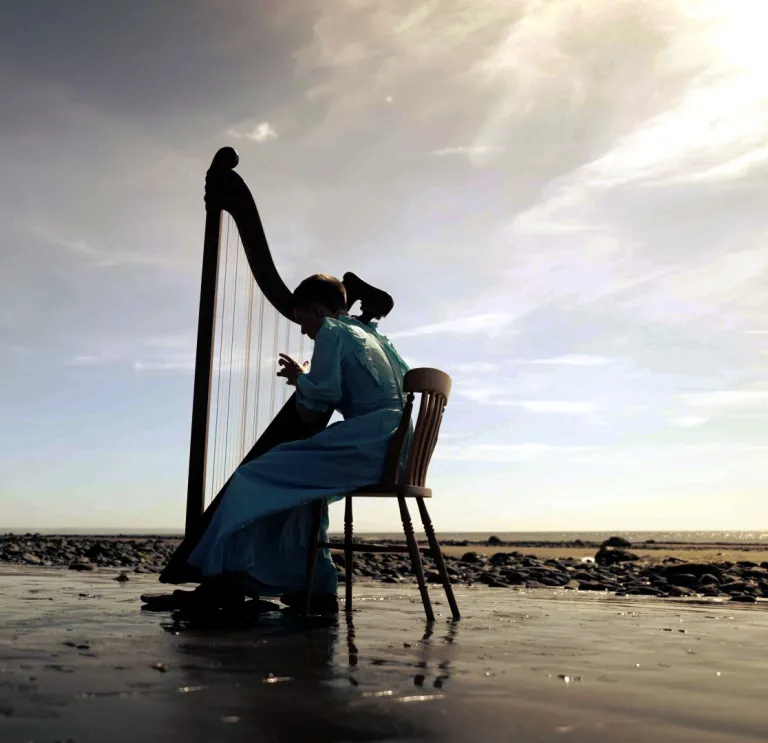 A young harp player wearing a long dress, sitting and playing a harp on a beautiful deserted beach