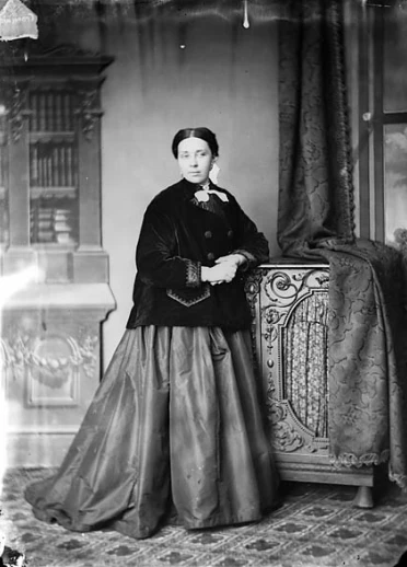 黒いボタン付きのジャケットとふんわりしたロングスカート、白いアクセサリーを身に着け、写真の右側を見つめている。