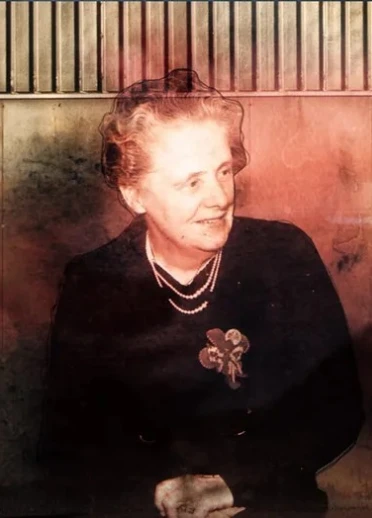 セピア調のホログラム映像のエリザベス。真珠のネックレスと花のブローチを着け、暗い服を身に着けている。