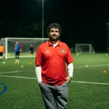 Club founder Delwyn Derrick standing on a football pitch while a game takes places behind him
