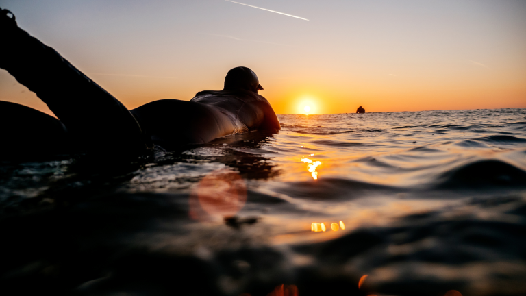 Surfer in wetsuit floating on the surface of the sea, silhouetted by the sunset on the horizon