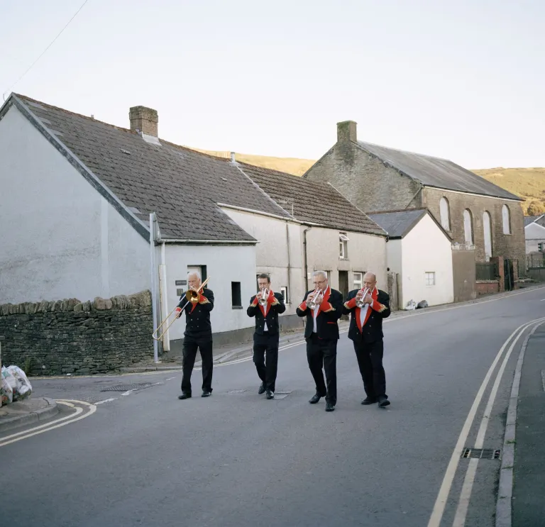 Four members of a traditional Welsh brass band playing their instruments, walking along a typical Welsh valleys street