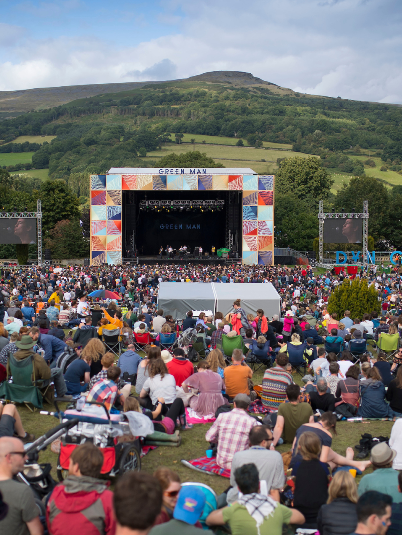 A crowd of people sat in a field looking towards an outdoor festival stage with a hill in the background
