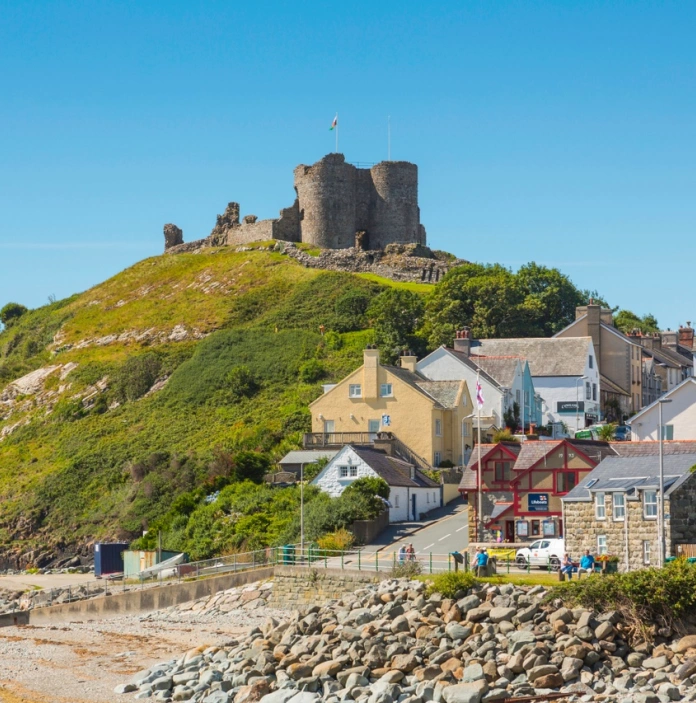 An external view of a large castle on top of a green hill, next to a beach
