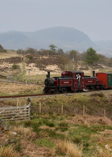 A red and green steam engine moving along a track in front of a mountainous backdrop