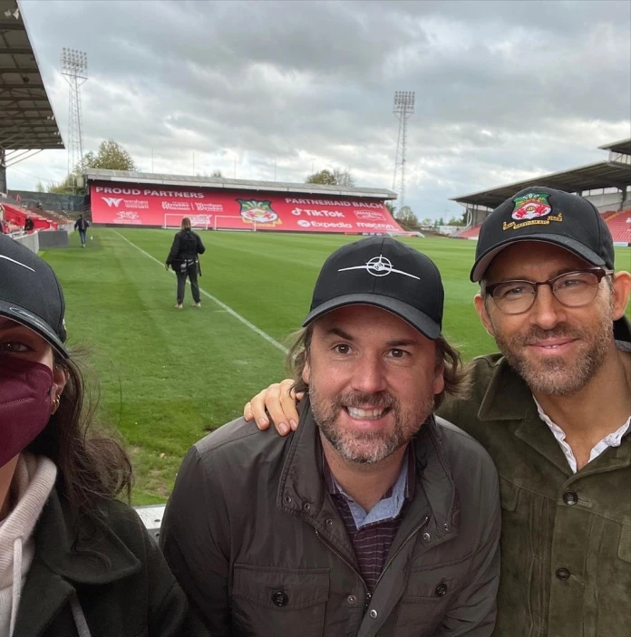 Three people on a football pitch smiling at the camera while filming takes place behind them