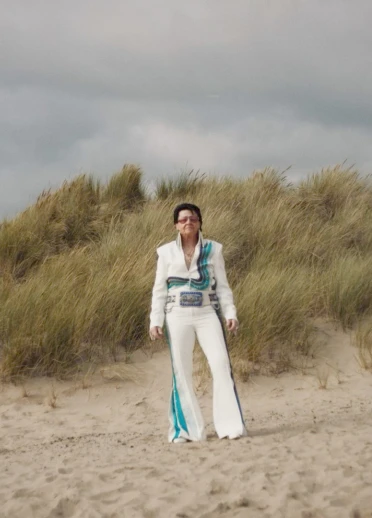 A female Elvis tribute artist dressed in a white embellished jumpsuit standing in sand dunes