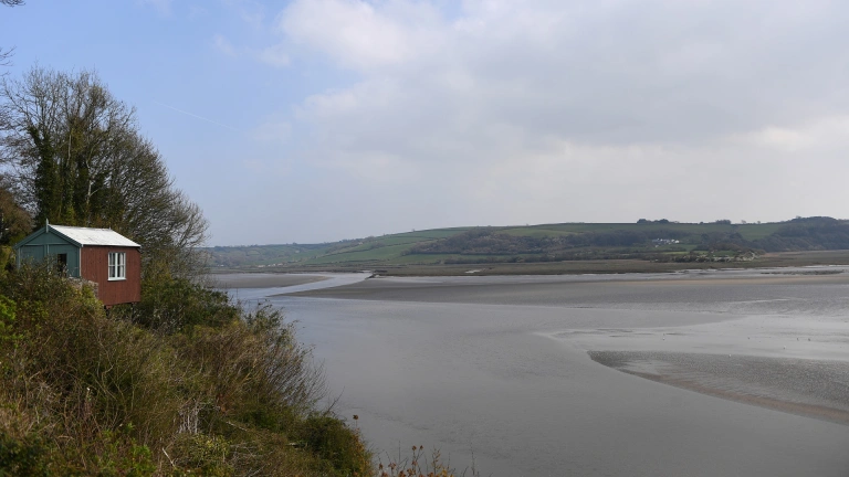A small pretty wooden shed perched on a forested cliff, overlooking a huge expansive estuary