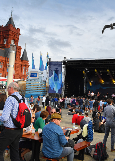 people sat and stood in crowd in foreground with outdoor stage in background