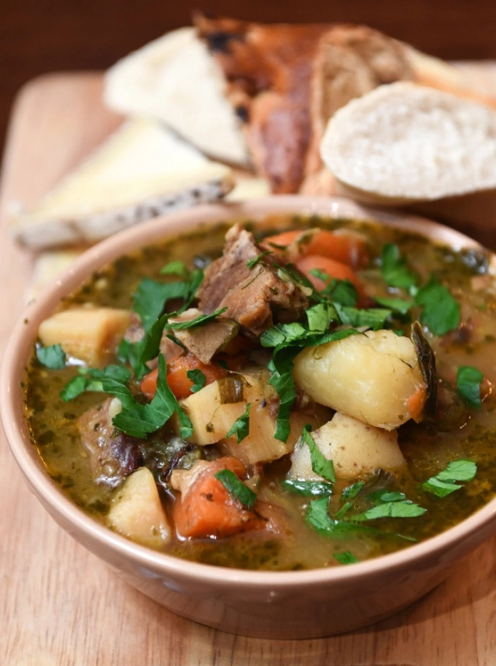 A bowl of cawl stew on a breadboard with bread 