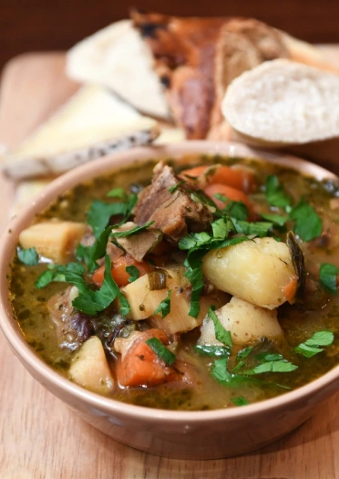 A bowl of cawl stew on a breadboard with bread 