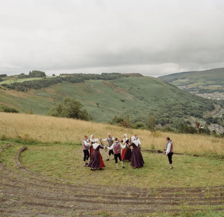 A group of dancers dressed in traditional outfits, dancing on a green hillside with valleys in the distance