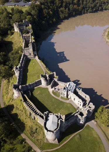 Aerial view of the ruins of an old castle located by a river and surrounded by green fields and trees.