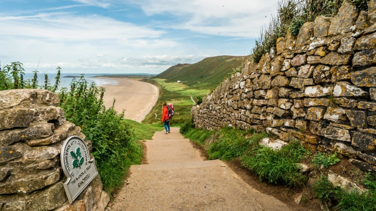 A person with a baby in a carrying backpack, standing on a walking path overlooking a faraway beach
