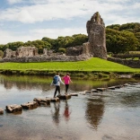 Horses and walkers in front of a ruined castle surrounded by green fields and water.